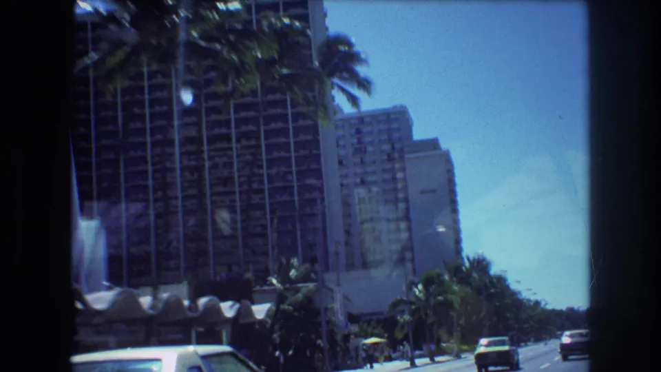 1977 Honolulu roadside view showing tall hotel buildings, palm trees, classic cars, and pedestrians along a sunny multi-lane street.