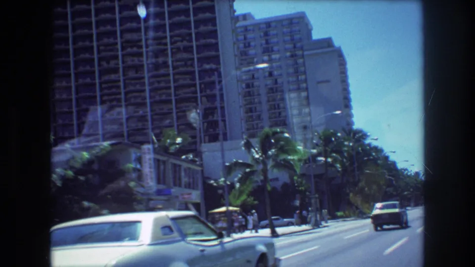 1977 Honolulu driving view with palm trees, high-rise hotel facades, a white coupe at the lower edge, and cars moving ahead on a broad street.