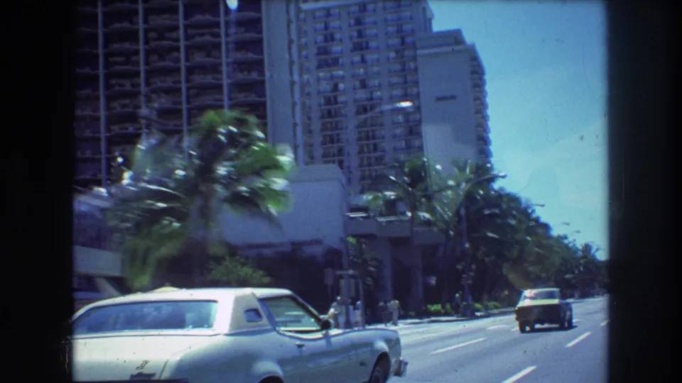 1977 Honolulu street scene from a moving car, with a white coupe in the foreground, hotel balconies above, palms along the curb, and traffic continuing down the road.