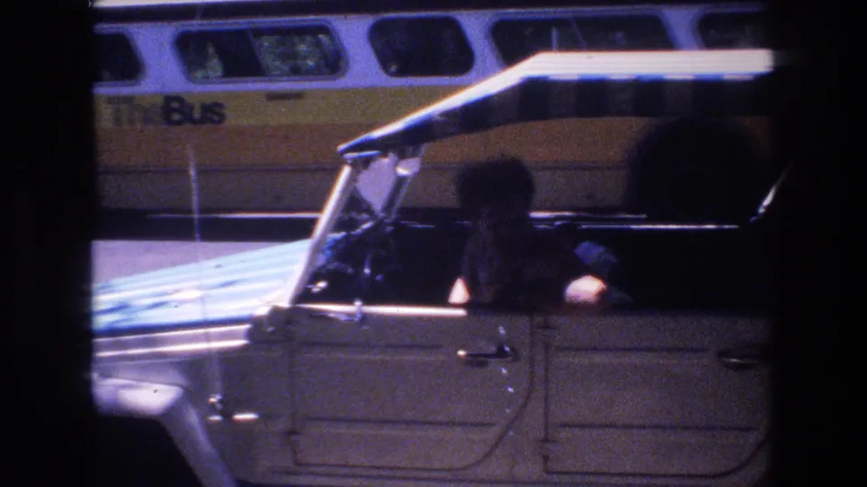 1977 Honolulu drive-by frame with a yellow bus outside and a person visible in silhouette inside or beside the filming vehicle, partially obscuring the street view.