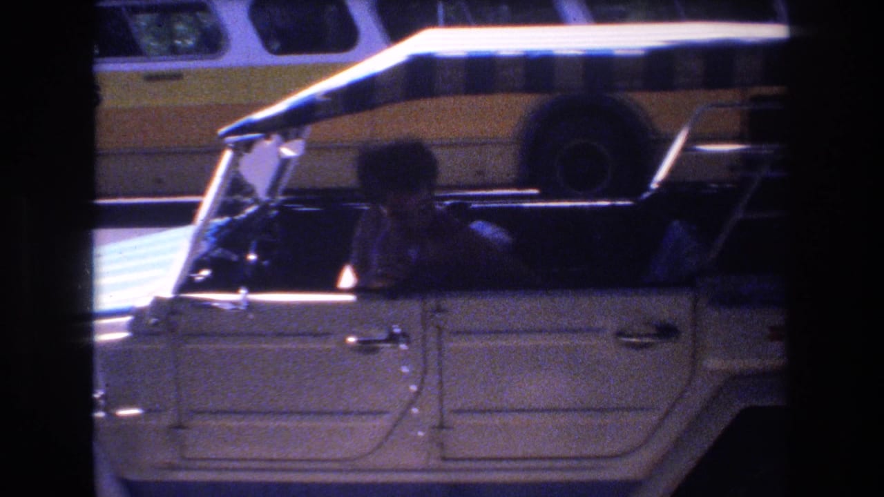 1977 Honolulu home-movie frame showing a person in the driver's seat of a white Volkswagen Thing with a striped canopy and a yellow-and-white bus in the background.
