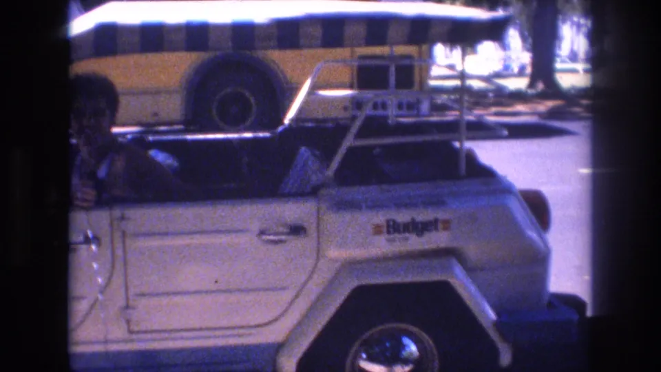 Super 8 frame from the Honolulu clip showing the side of the white Volkswagen Thing, a partially visible driver, the striped canopy, and the Budget marking on the vehicle side or background area.