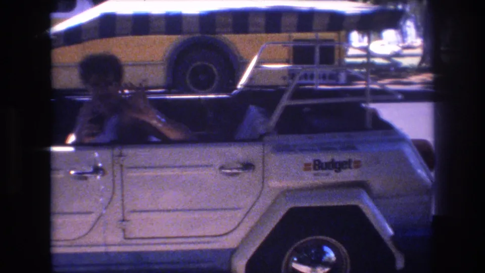 1977 Honolulu frame showing the driver gesturing toward the camera from a white Volkswagen Thing, with the striped canopy and a yellow-and-white bus in the background.