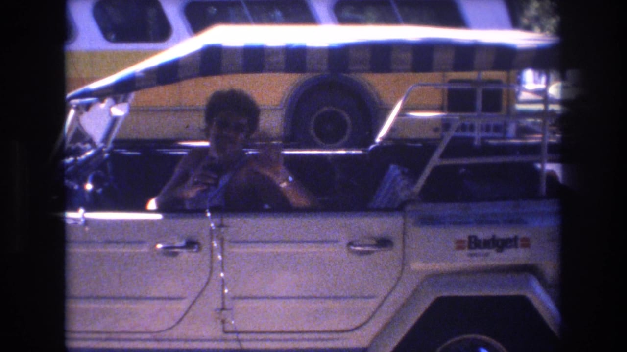In 1977 Honolulu home-movie footage, a man sits in the driver's seat of a white open-top car with a striped canopy while a yellow-and-white bus passes or stands behind him.
