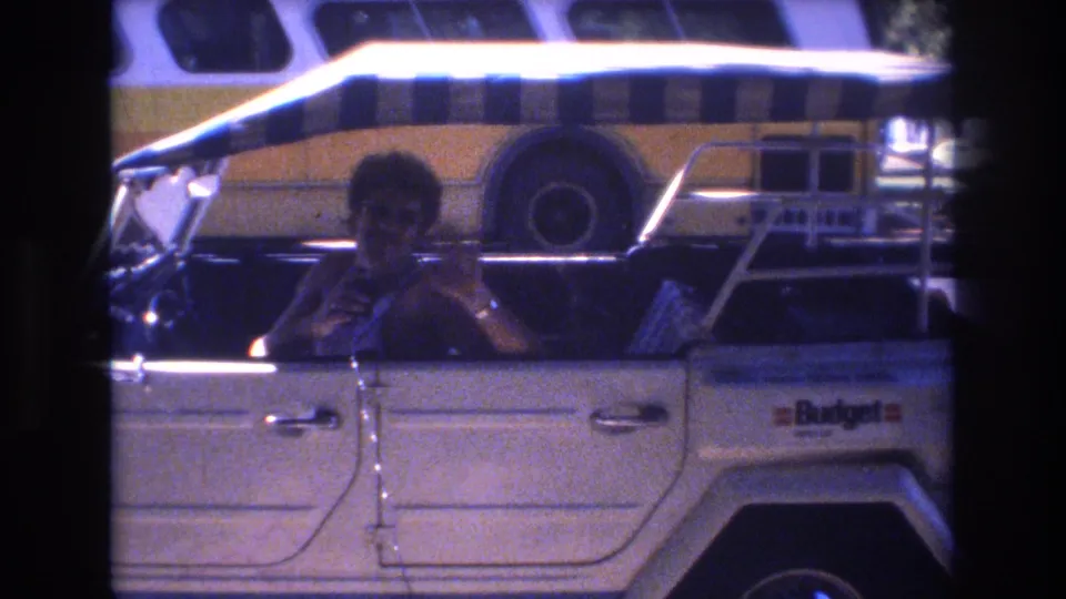In 1977 Honolulu home-movie footage, a man sits in the driver's seat of a white open-top car with a striped canopy while a yellow-and-white bus passes or stands behind him.