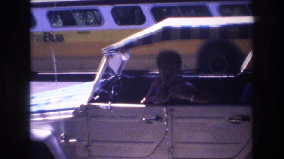 A darker frame shows the man partly in shadow inside the white open-top car, with the canopy crossing the frame and bus windows visible behind the vehicle.