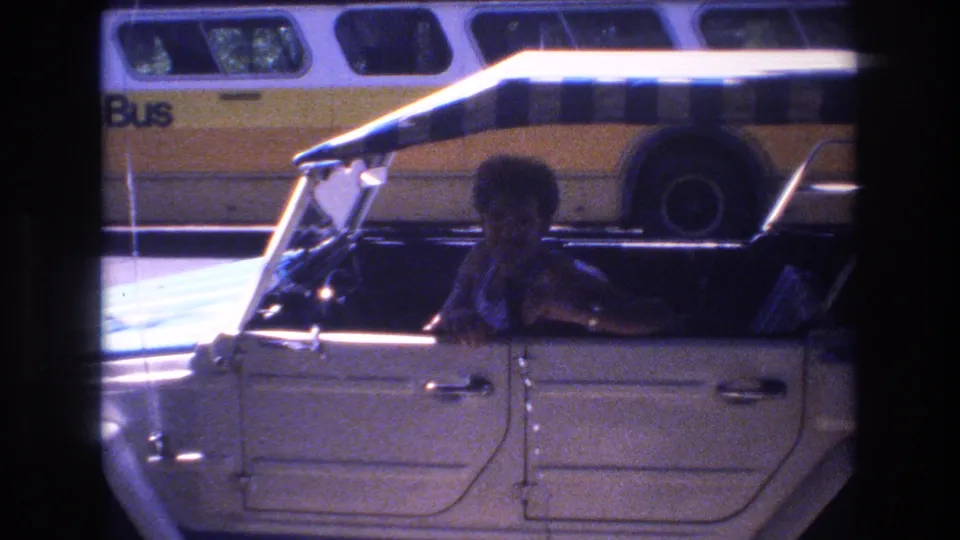 The man appears to gesture or speak from the driver's seat of the white open-top car, with the striped canopy and yellow-and-white bus still visible behind him in the 1977 Honolulu footage.