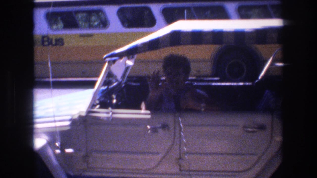 1977 Honolulu home-movie frame showing a woman in or beside a light-colored car, with a yellow bus visible in the background and dark 8mm frame edges.