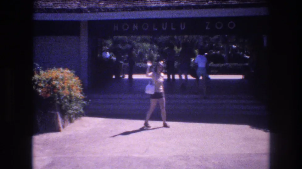 1977 Honolulu frame showing a woman in a light top and shorts waving in the paved forecourt outside the Honolulu Zoo entrance, with visitors and greenery behind her.