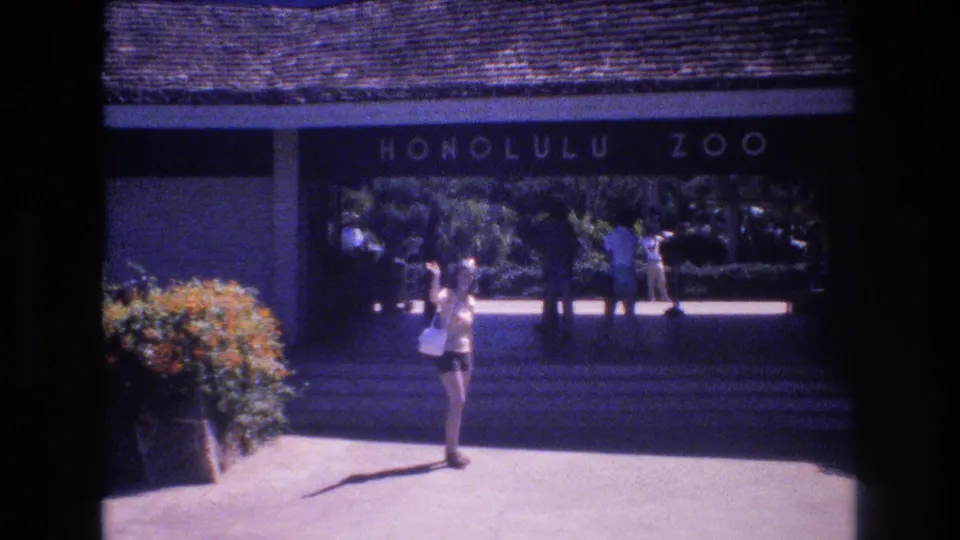 1977 Honolulu Zoo entrance frame with the woman centered on the pavement, arm raised toward the camera, and several visitors visible under the shaded walkway.