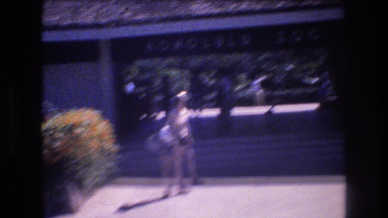 A distant person in light-colored clothing stands on a walkway near dark building shade and a rounded shrub in 1977 Honolulu home-movie footage.