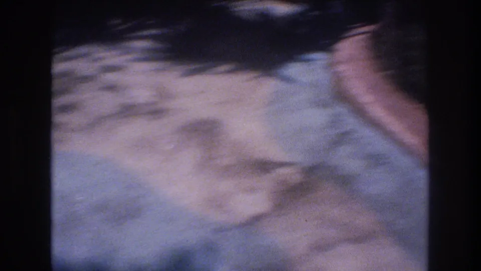 A blurred 1977 Honolulu 8mm frame shows a paved area with strong shadows and partial motion at the right edge, likely from handheld camera movement.