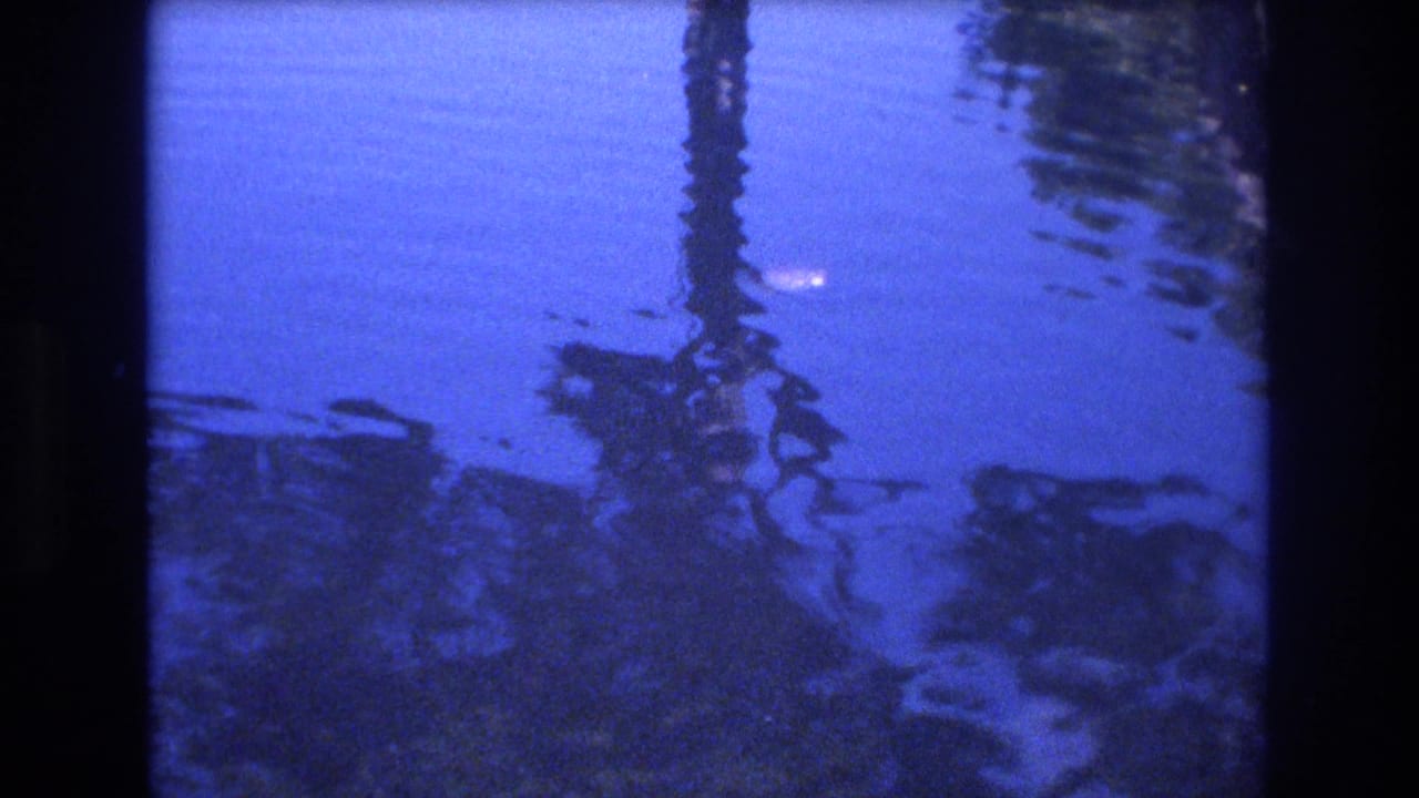 1977 Honolulu 8mm home-movie frame showing blue-tinted water with dark reflected vertical forms and subtle surface ripples.