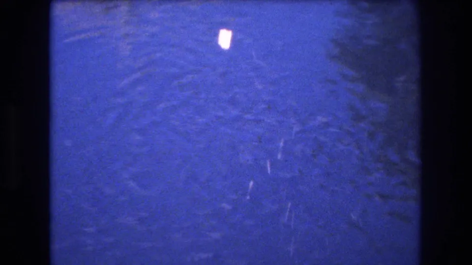 1977 Honolulu 8mm home-movie frame showing a small bright piece, likely bread, hitting blue water and spreading ripples.