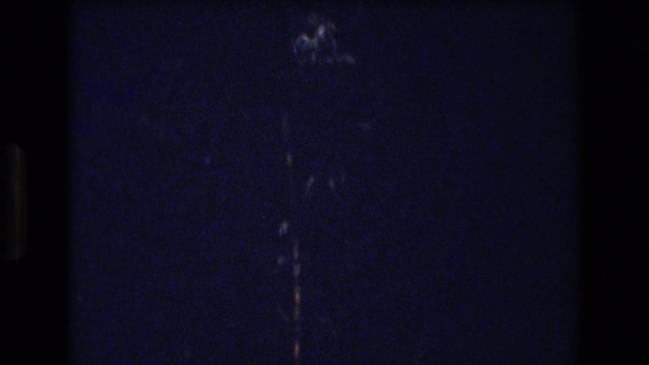Dark grainy frame with a faint vertical light mark near the center.
