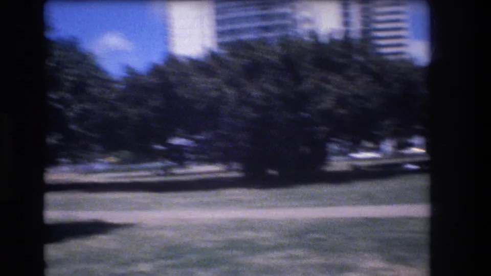 Handheld 8mm Honolulu frame showing trees, grass, a paved edge, and tall city buildings beyond the park landscaping.