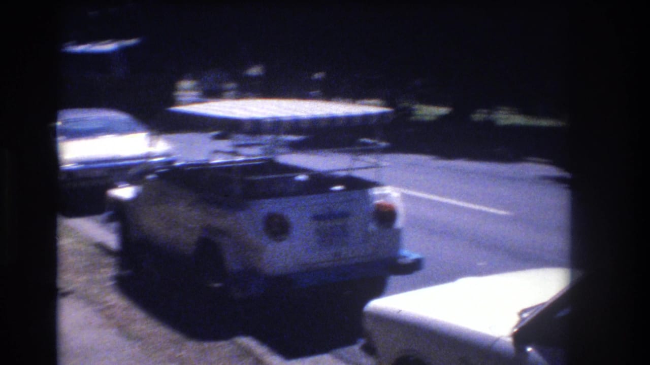 In 1977 Honolulu, a white Volkswagen Thing with a striped canopy is parked curbside while a large yellow bus moves through the background.
