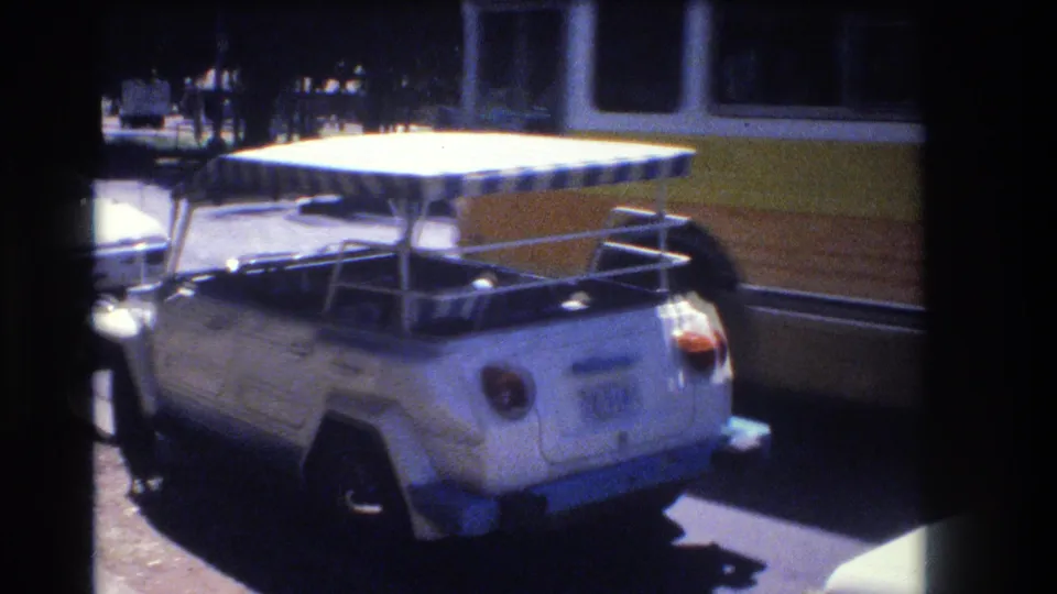 A rear three-quarter view shows the white Volkswagen Thing and its blue-and-white canopy, with a yellow bus close behind on the street in Honolulu, 1977.