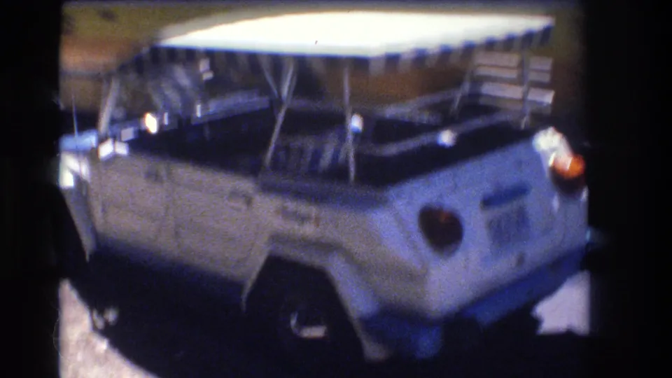 The camera moves closer to the rear of the white Volkswagen Thing, emphasizing the striped canopy, spare tire, and soft 8mm color shift in the 1977 Honolulu street scene.