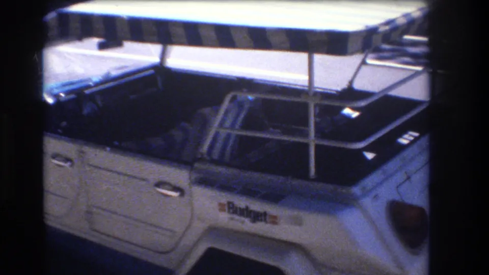 A side-rear angle of the parked white Volkswagen Thing shows the open seating area, canopy supports, chrome rails, and small side markings in the 1977 Honolulu footage.