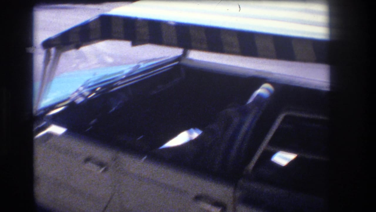1977 Honolulu home-movie frame looking past the side of a car, with a dark window, roof edge, windshield reflection, and pale building or awning above.