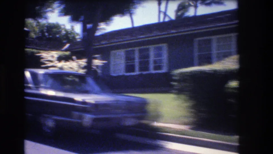 1977 Honolulu home-movie frame with a car in the foreground passing a single-story residence, clipped hedges, lawn, and palm trees.