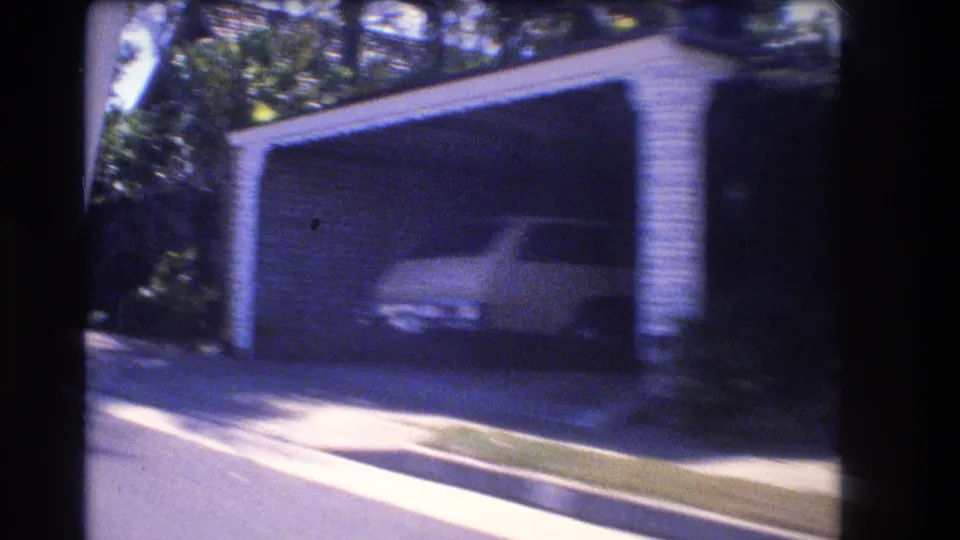 1977 Honolulu street-side view of an open garage or carport with a parked vehicle, driveway, curb, and surrounding tropical greenery.