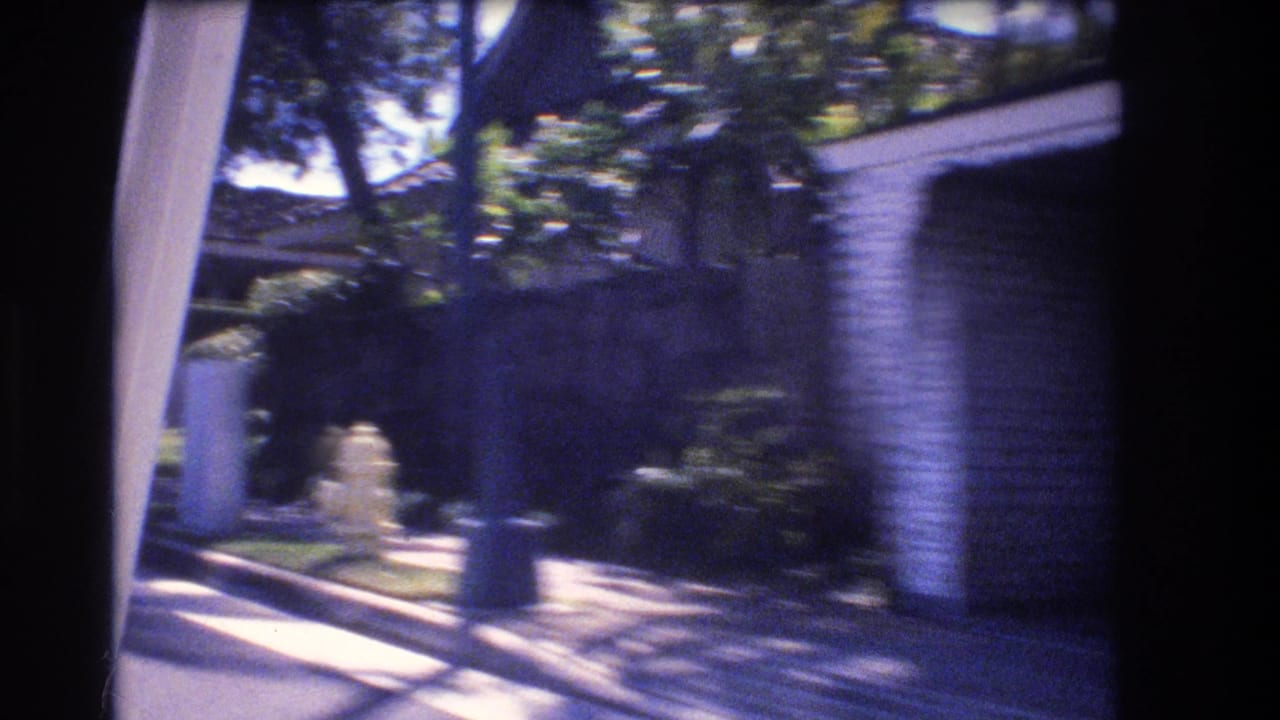 1977 Honolulu home-movie frame from a moving car showing a sunlit residential street with a low house, hedges, sidewalk, and a blurred possible person near the curb.