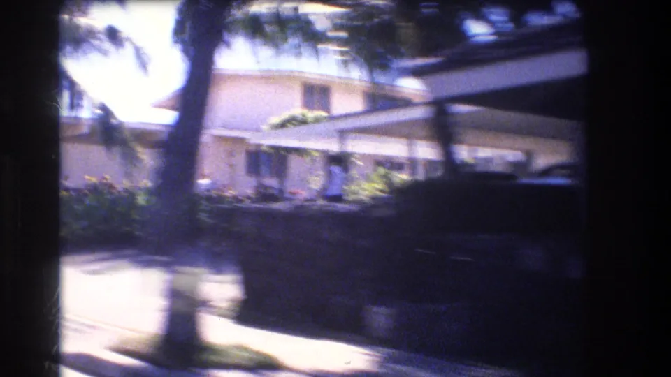 1977 Honolulu home-movie frame showing a residential house with a carport or garage area, palm trees, hedge planting, and deep shade from the passing vehicle view.