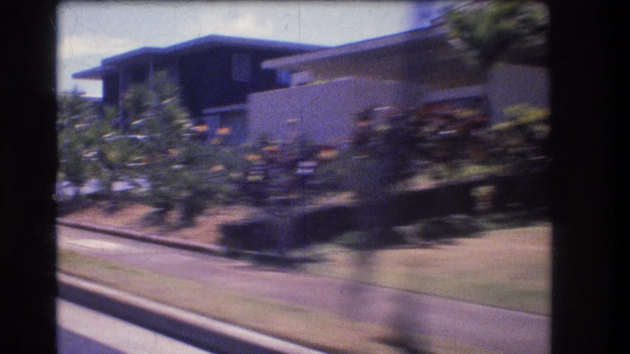 1977 Honolulu home-movie frame from a moving vehicle passing a flat-roofed suburban house with sidewalk, lawn, hedges, and tropical landscaping.