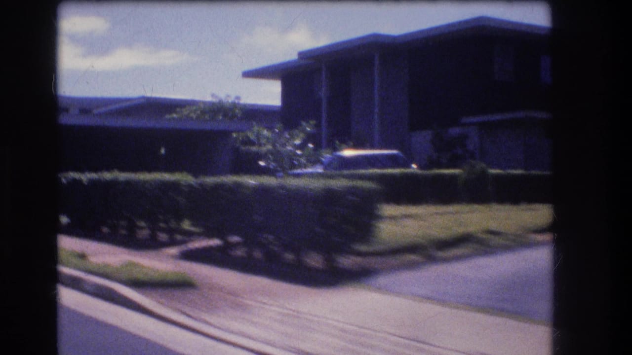 Residential corner with hedges, driveway, flat-roofed house, and a parked car partly visible.