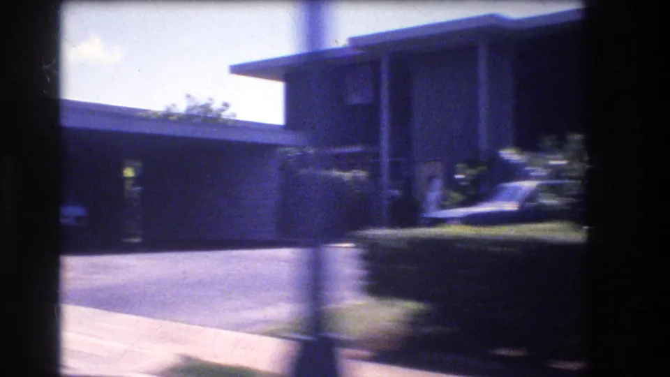 1977 Honolulu home-movie frame passing a flat-roofed house with a driveway or carport area, low wall, hedge, sidewalk, and foreground motion blur.