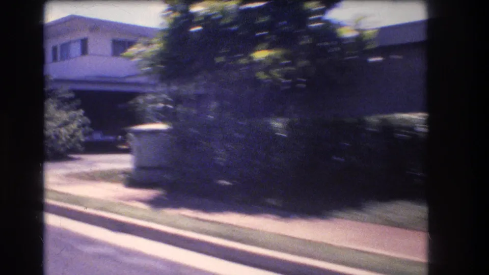 1977 Honolulu residential street frame showing a house partly hidden by tropical trees and hedges, with a curb, sidewalk, driveway, and strong motion blur.