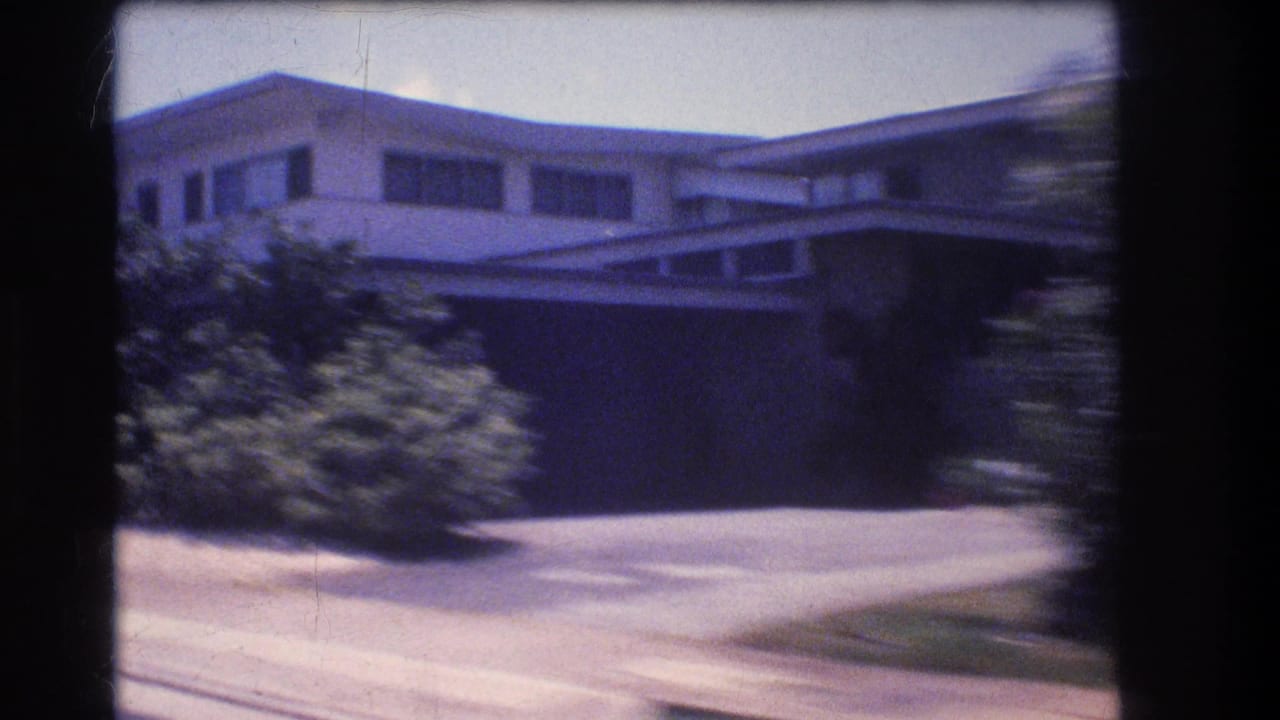 1977 Honolulu 8mm frame showing a low modernist house with broad windows, front bushes, sidewalk edge, and motion blur from a passing vehicle.
