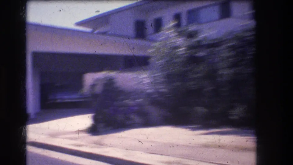 1977 Honolulu home-movie frame showing an attached garage, driveway, low house roofline, and foreground shrubs blurred by vehicle movement.