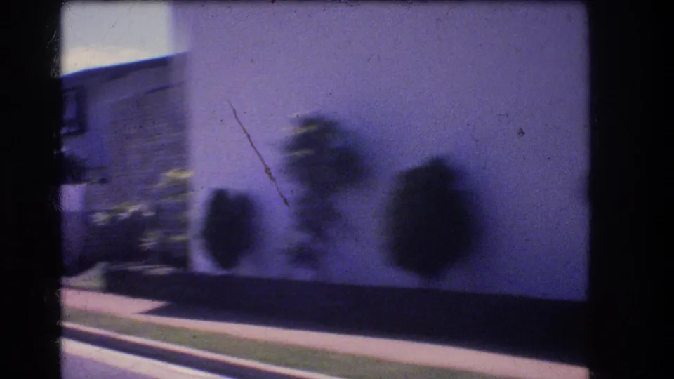 1977 Honolulu 8mm frame with a pale residential wall, clipped shrubs, sidewalk edge, and strong blur from the moving vehicle viewpoint.