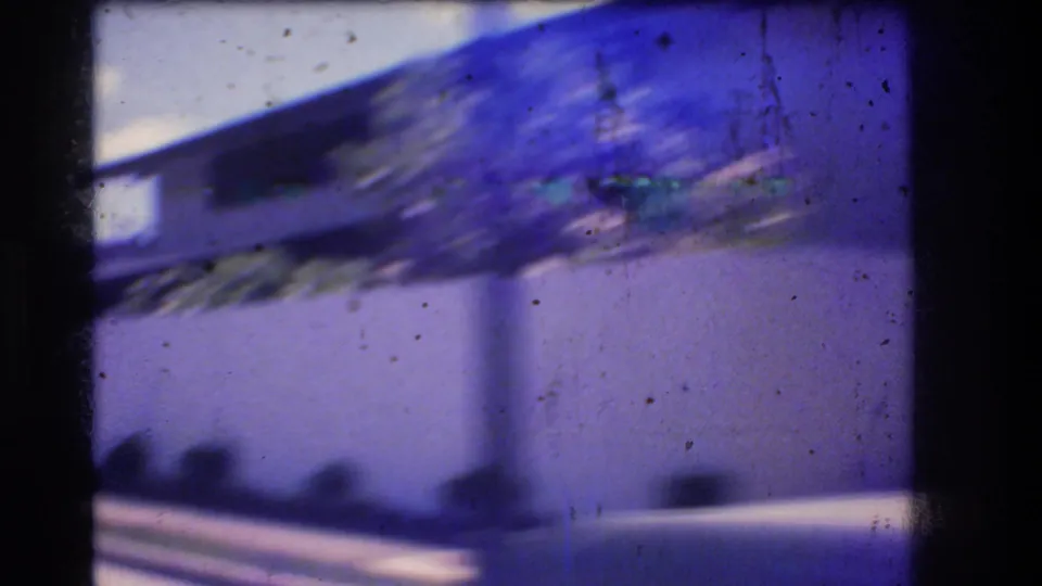 1977 Honolulu 8mm frame showing a blurred modernist roofline above a pale wall, roadside plantings, and visible dust and scratches on the film.