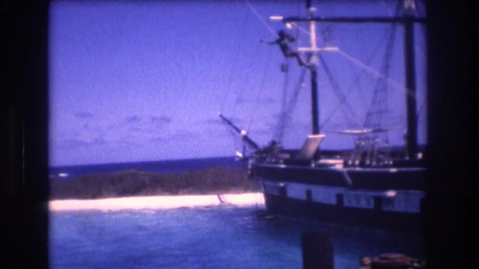 A 1977 Honolulu Oahu home-movie frame shows a sail-rigged boat near blue water, a pale beach, and a low tropical shoreline in soft Super 8 color.