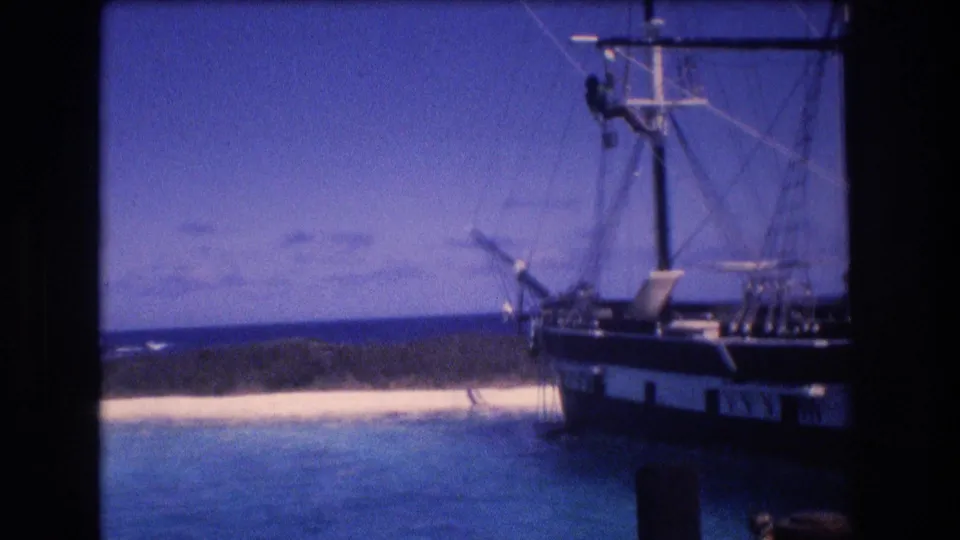 The closing 1977 Honolulu-area frame holds on the rigging and hull of a sail-rigged boat near a bright beach and blue coastal water.