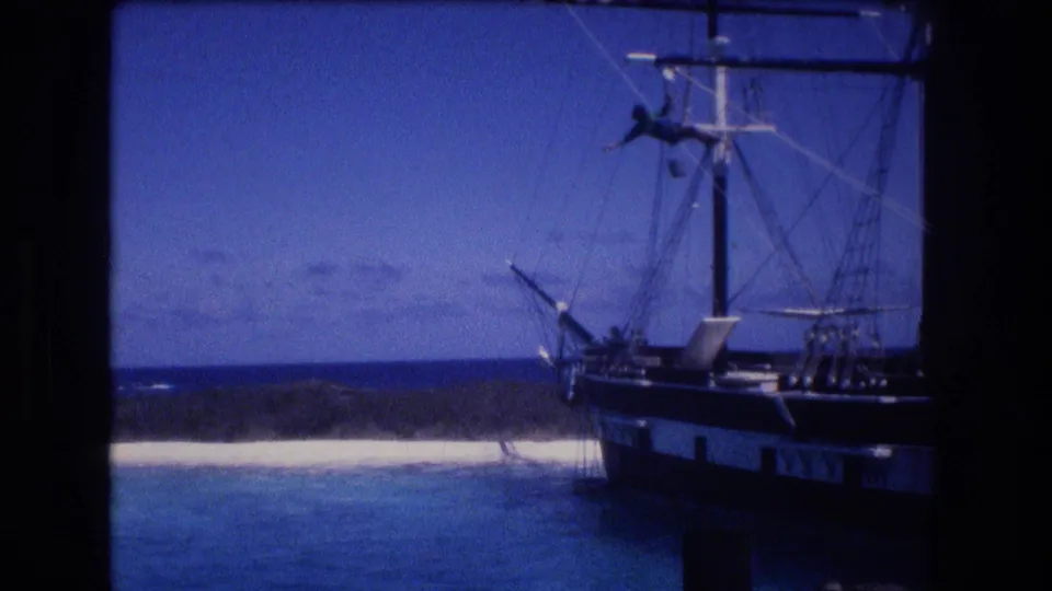 1977 Honolulu, Oahu frame showing a crew member silhouetted on a sailing vessel mast, with the dark hull, rigging, blue bay, and pale shoreline below.