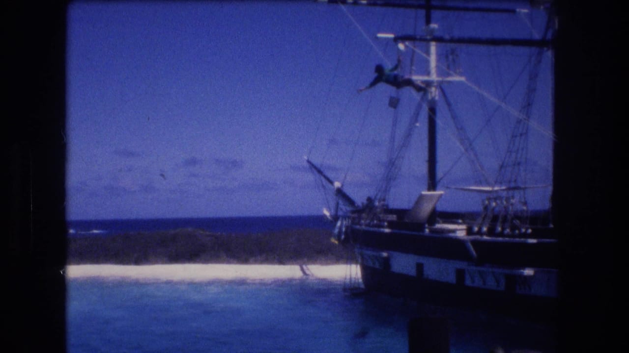 1977 8mm home-movie frame from Honolulu Oahu showing a dark-hulled sailing ship near blue water, pale beach, and low green coastline, with a person visible on the rigging.