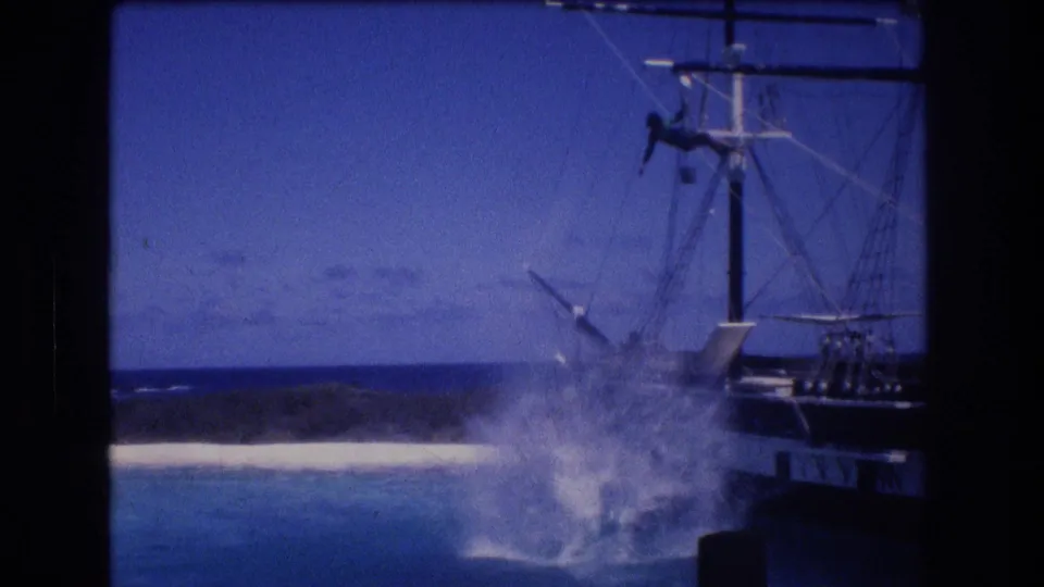 1977 8mm home-movie frame from Honolulu Oahu showing a tall splash in the ocean beside a dark-hulled sailing ship, with rigging, beach, coastline, and sky visible.