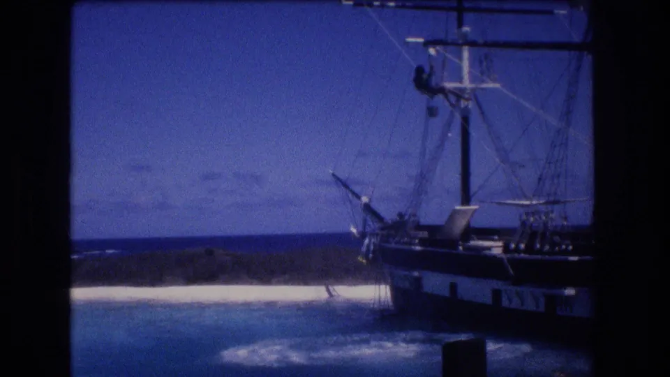 1977 8mm home-movie frame from Honolulu Oahu showing foam and ripples where the dolphin entered the water beside the sailing ship, with rigging, shoreline, and sky behind.