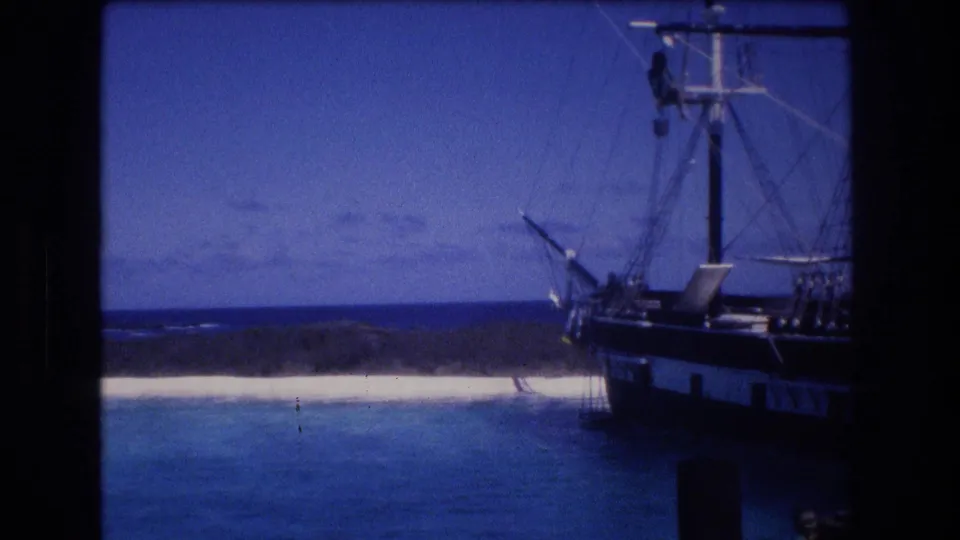 1977 Honolulu Oahu home-movie frame showing the tall rigging of a docked sailing vessel, a small figure aloft, calm water, pale beach, and blue horizon under soft twilight exposure.