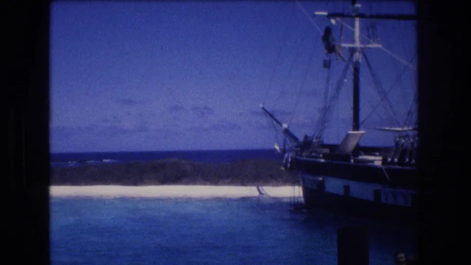 1977 Honolulu Oahu home-movie frame with a sailing vessel and rigging at the right side, blue water in the foreground, pale shoreline, and a distant ocean horizon.