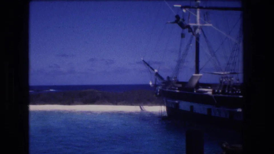 1977 Honolulu-area frame of a docked or near-shore sailing ship, with a small person visible on the mast over turquoise water and a pale beach.