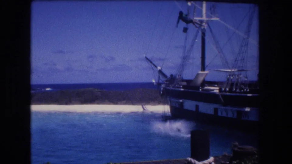 1977 Honolulu, Oahu home-movie frame with a large splash beside the ship after the dolphin movement, with beach, shoreline, and rigging visible in the background.