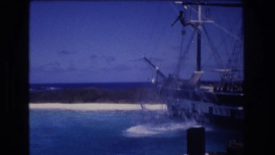 1977 Honolulu, Oahu frame showing a broad splash on turquoise water beside the rigged ship, with a person still visible high on the mast against the blue sky.