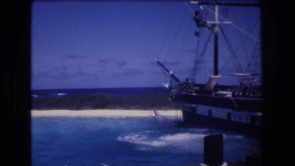 1977 Honolulu, Oahu home-movie frame showing a large moored sailing ship near turquoise water, white sand, low shoreline, and blue sky.