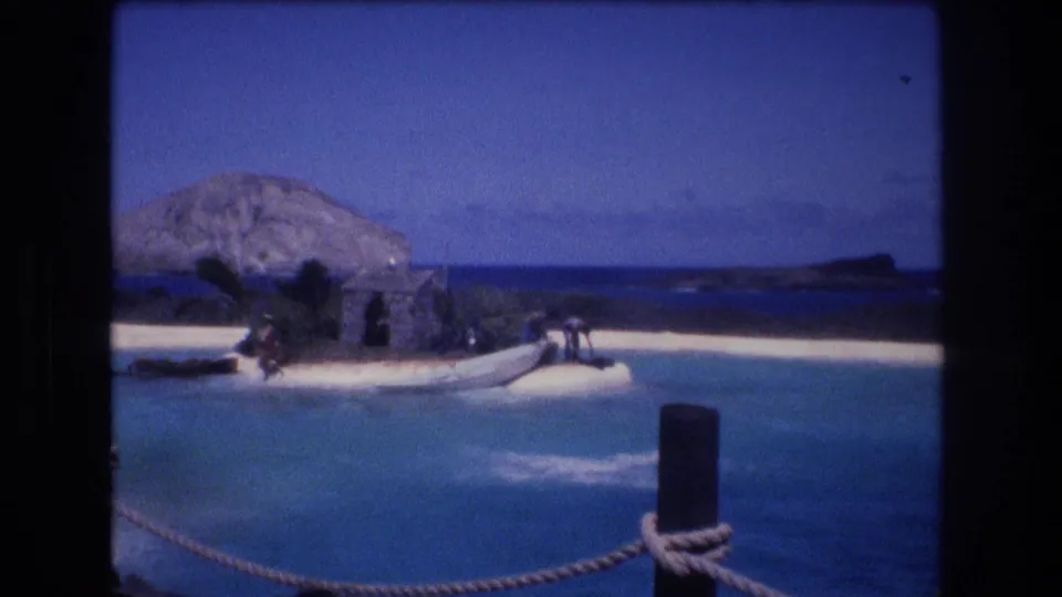 1977 Honolulu, Oahu 8mm frame showing the aftermath of a marine animal jump in a blue lagoon, with a sandy island set, boat, and distant people visible.